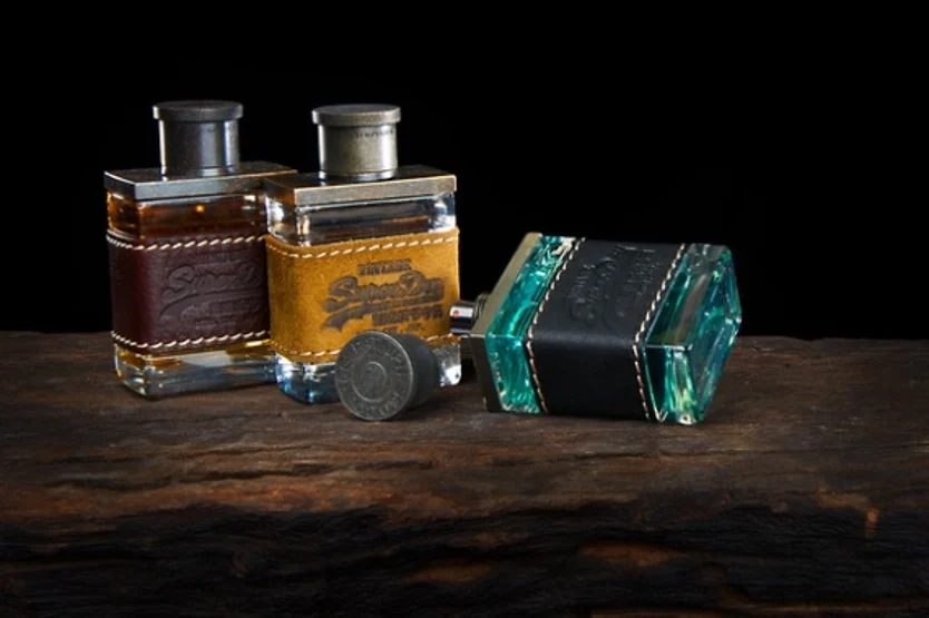 Three Superdry fragrances in leather-bound flacons with knurled metal caps, on weathered wood